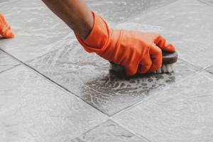Hand with orange glove scrubs the tile floor with a brush.